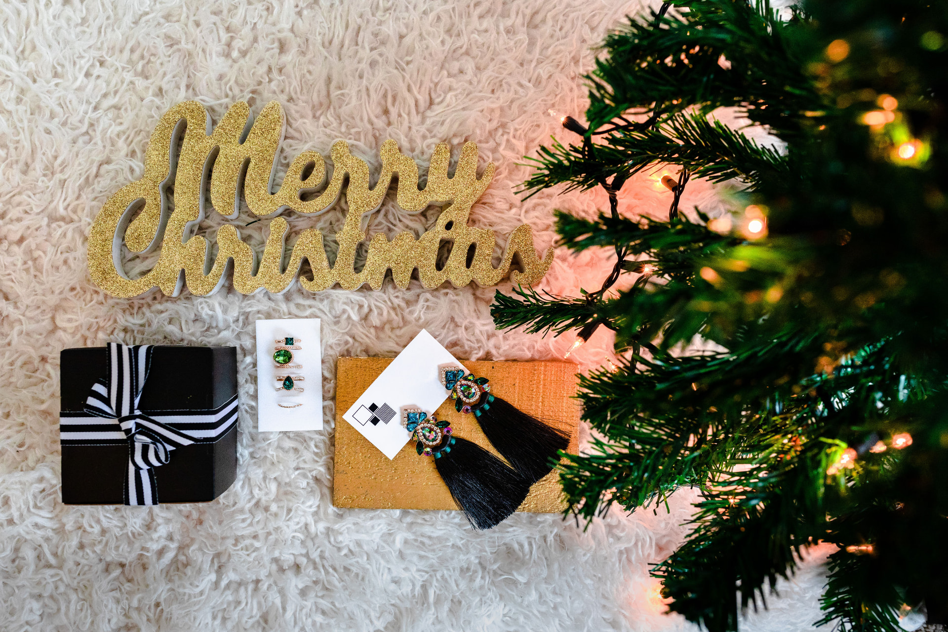 Festive holiday flat lay with gold Merry Christmas sign, jewelry and gift box beside a lit tree